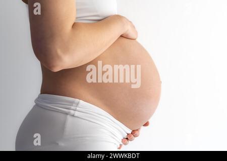 Beschreibung: Mittelteil der stehenden Frau, die sanft ihre sehr runde Schwangere hält. Seitenansicht. Weißer Hintergrund. Brillante Aufnahme. Stockfoto