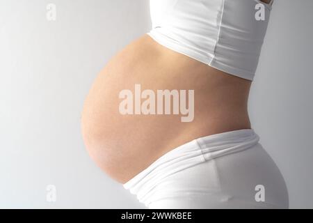 Beschreibung: Mittelteil einer nicht erkennbaren stehenden Mutter in weißen Tüchern mit sehr rundem Schwangerschaftsbauch. Seitenansicht. Weißer Hintergrund. Helles Licht Stockfoto