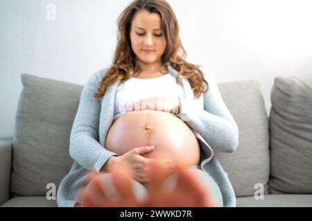 Beschreibung: Frontalansicht einer glücklichen Frau, die auf dem Sofa sitzt und sanft ihren Bauch hält, in Erwartung eines Babys im letzten Stadium der Schwangerschaft. Pregnanc Stockfoto