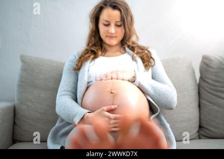 Beschreibung: Frontalansicht einer glücklichen Frau, die auf dem Sofa sitzt und sanft ihren Bauch hält, in Erwartung eines Babys im letzten Stadium der Schwangerschaft. Pregnanc Stockfoto
