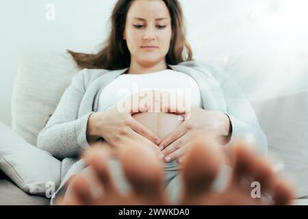 Beschreibung: Frontalansicht einer Frau, die in Erwartung ihres Babys in den letzten Monaten der Schwangerschaft auf dem Sofa liegend, sanft ihren Bauch hält. Schwangerschaftsdrittstrumpf Stockfoto