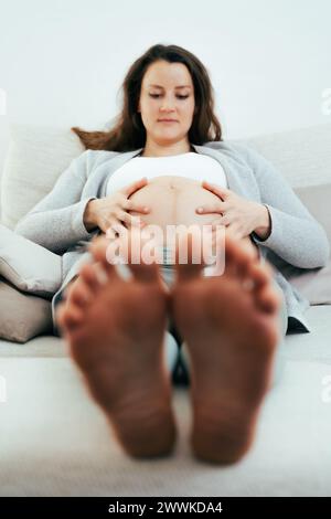 Beschreibung: Frontalansicht einer Frau, die auf dem Sofa liegt und sanft ihren Bauch hält, in glücklicher Erwartung ihres Babys in den letzten Monaten der Schwangerschaft. Schwangerschaft durch Stockfoto
