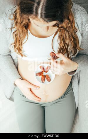 Beschreibung: Draufsicht einer Frau, die in den letzten Monaten der Schwangerschaft auf dem Sofa sitzt und Daddles isst, während sie ihren Bauch sanft hält. Schwangerschaft dritte tr Stockfoto