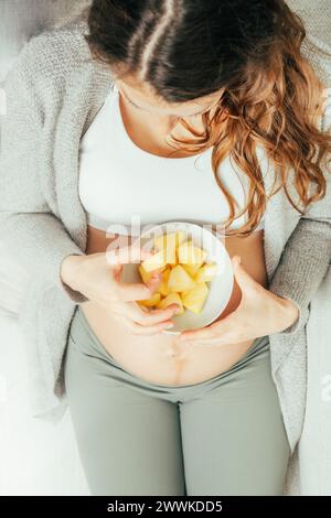 Beschreibung: Draufsicht einer Frau, die in den letzten Monaten der Schwangerschaft auf dem Sofa sitzt und Ananas-Stücke isst, während sie ihren Bauch sanft hält. Schwangerschaft Th Stockfoto