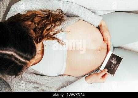 Beschreibung: Draufsicht einer Frau, die auf einem Sofa sitzt und ein Ultraschallbild ihres Babys während der Schwangerschaft ansieht. Schwangerschaft drittes Trimenon - Woche 34. Oben Stockfoto