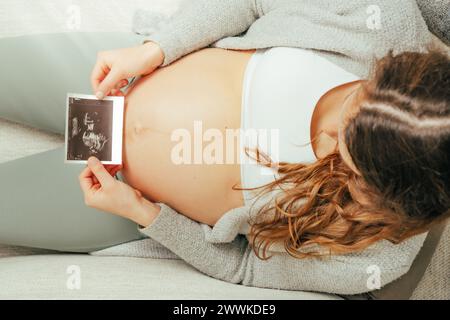 Beschreibung: Draufsicht einer Frau, die auf einem Sofa sitzt und ein Ultraschallbild ihres Babys im letzten Stadium der Schwangerschaft ansieht. Schwangerschaft drittes Trimeste Stockfoto