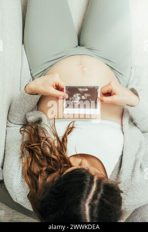 Beschreibung: Draufsicht einer Frau, die auf einem Sofa sitzt und ein Ultraschallbild ihres Babys im letzten Stadium der Schwangerschaft ansieht. Schwangerschaft drittes Trimeste Stockfoto