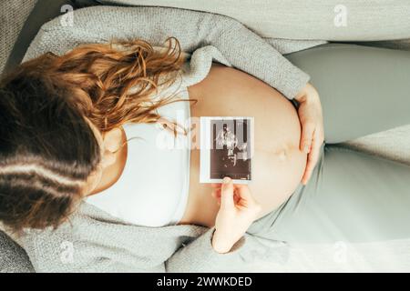 Beschreibung: Draufsicht einer Frau, die auf einem Sofa sitzt und ein Ultraschallbild ihres Babys im letzten Stadium der Schwangerschaft ansieht. Schwangerschaft drittes Trimeste Stockfoto