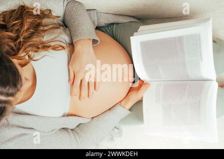 Beschreibung: Draufsicht einer Frau, die auf einem Sofa sitzt und im letzten Stadium der Schwangerschaft ein Buch liest. Schwangerschaft drittes Trimenon - Woche 34. Draufsicht. Sonnig Stockfoto