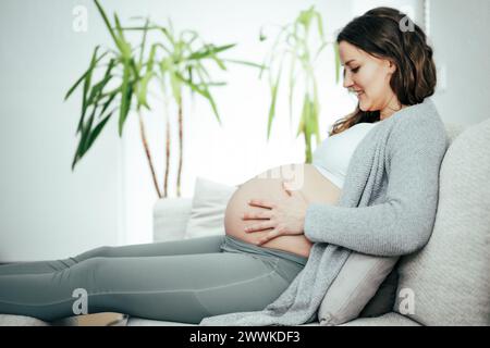 Beschreibung: Seitenansicht einer Frau, die auf einem Sofa sitzt und mit ihrem Baby in ihrem Bauch während der letzten Phase der Schwangerschaft spielt. Schwangerschaftsdritttrimest Stockfoto