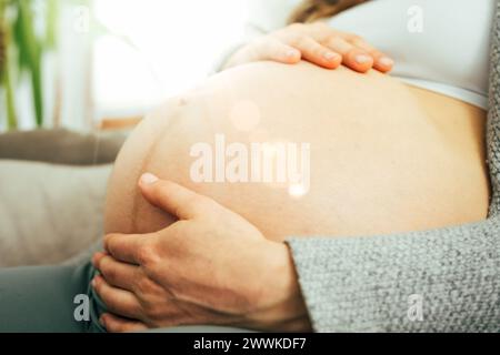 Beschreibung: Nahaufnahme einer Frau, die im letzten Stadium der Schwangerschaft auf einem Sofa sitzt und sanft ihren Bauch hält. Schwangerschaft drittes Trimenon - Woche 34. Seite VI Stockfoto