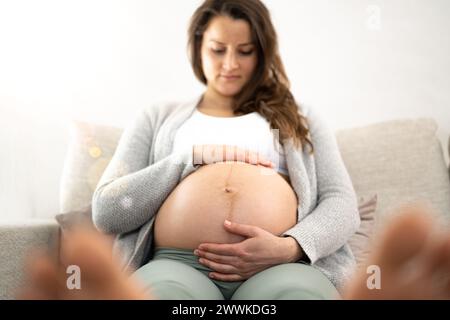 Beschreibung: Frontalansicht einer Frau, die auf einem Sofa sitzt und in Erwartung des Babys während des letzten Schwangerschaftsstadiums sanft ihren Bauch hält. Schwangerschaft Stockfoto