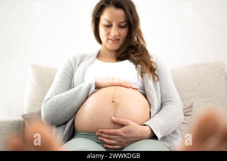 Beschreibung: Frontalansicht einer lächelnden Frau, die auf einem Sofa sitzt und sanft ihren Bauch hält, in Erwartung des Babys während des letzten Schwangerschaftsstadiums. P Stockfoto