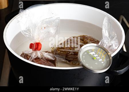 Beschreibung: Zubereitung von rohem Rinderfiletsteak für ein Sous-Vide-Bad in einem Topf mit Thermometer auf dem Küchenherd, von der Seite fotografiert. Stockfoto