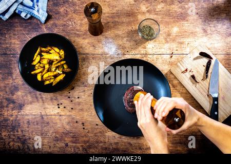 Beschreibung: Man würzt ein hausgemachtes Rinderfiletsteak, serviert auf einem schwarzen Teller neben einer schwarzen Schüssel mit Kartoffelspalten, Pfefferstreuer, Rosmarin und einem w Stockfoto