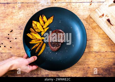 Beschreibung: Hand eines Mannes, der ein hausgemachtes Rinderfiletsteak mit knusprigen goldbraunen Kartoffelkeilen hält, serviert auf einem schwarzen Teller neben einem Holzbrett Stockfoto