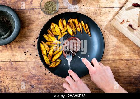 Beschreibung: Ein Mann schneidet ein hausgemachtes Rinderfiletsteak mit knusprigen goldbraunen Kartoffelkeilen, serviert auf einem schwarzen Teller neben einem Steinmörtel Stockfoto