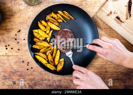 Beschreibung: Ein Mann schneidet ein hausgemachtes Rinderfiletsteak mit knusprigen goldbraunen Kartoffelspalten, serviert auf einem schwarzen Teller neben Rosmarin und einem Holzbrett Stockfoto