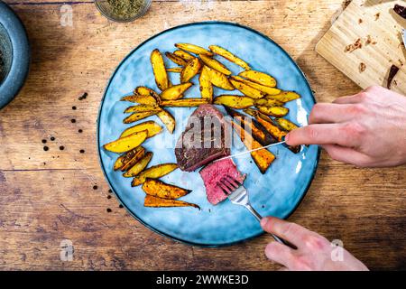 Beschreibung: Ein Mann schneidet ein hausgemachtes mittelgroßes Rinderfilet mit knusprigen goldbraunen Kartoffelspalten auf einem blauen Teller neben einem Steinmörtel, Ro Stockfoto