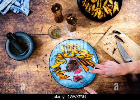 Beschreibung: Ein Mann hält ein hausgemachtes Filetsteak mit knusprigen goldbraunen Kartoffelspalten, serviert auf einem blauen Teller neben einer schwarzen Schüssel Stockfoto