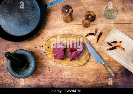 Beschreibung: Zubereitung von rohem Rinderfiletsteak auf Holzbrett mit Stahlpfanne, Mörtelsalz und Pfeffershakerand Rosmarin und kühlem Schneidebrett p Stockfoto