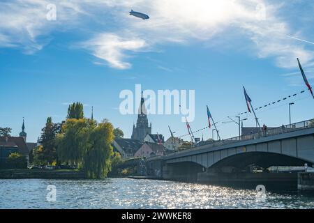 Beschreibung: Zeppelin fliegt an einem sonnigen Tag über die Stadt mit Touristen, die über eine Brücke auf der Steigenberger Halbinsel im Vordergrund laufen. Nachteile Stockfoto