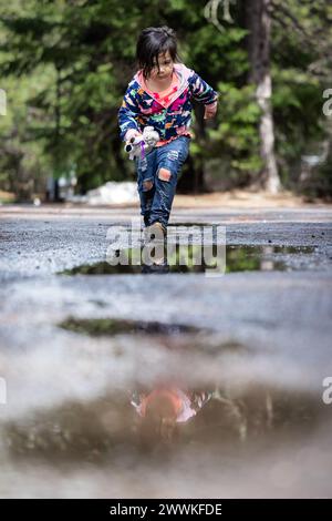 Ein junges Mädchen läuft auf einer nassen Straße, mit ihrem Spiegelbild im Wasser. Die Szene ist friedlich und ruhig, mit dem Mädchen, das ihre Zeit im Freien genießt Stockfoto