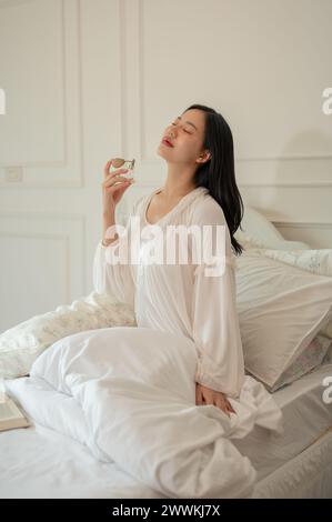 Eine attraktive asiatische Frau im langen Pyjama trägt Parfüm auf, nachdem sie auf ihrem Bett aufgewacht ist. Wohnleben und Lebensstil Stockfoto