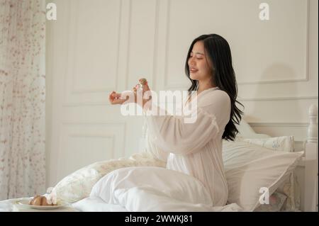 Eine attraktive asiatische Frau im langen Pyjama trägt Parfüm auf, nachdem sie auf ihrem Bett aufgewacht ist. Wohnleben und Lebensstil Stockfoto
