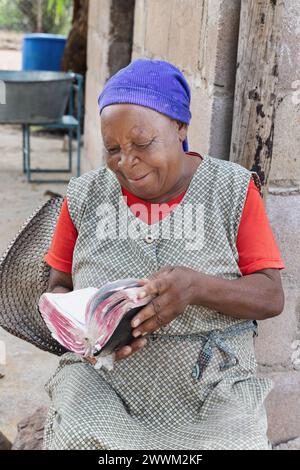 Glückliche christlich-afrikanische alte Frau, die die Bibel liest, in ihrem Garten sitzt, in einer Hütte im Hintergrund Stockfoto