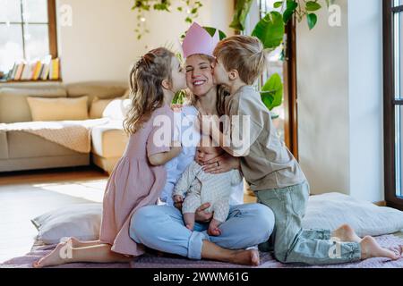 Mutter nach der Geburt als Königin. Tochter und Sohn küssen Mutter mit Papierkrone auf dem Kopf. Bewunderung, Liebe zu Müttern, Vorstellung vom Muttertag und Stockfoto