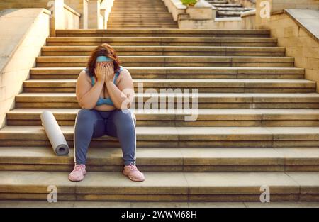 Traurige müde junge übergewichtige Frau mit Sportmatte, die auf Steintreppen sitzt und weint Stockfoto