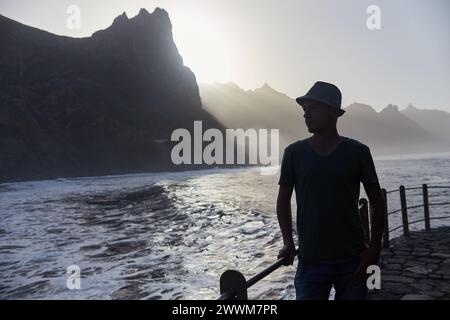 Mann, der an einem Sandstrand neben dem riesigen Meer steht, die Wellen, die sanft am Ufer unter einem Sonnenuntergang schwimmen Stockfoto