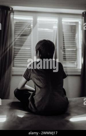 Junge auf dem Bett mit Licht, das durch Jalousien in der Pandemie leuchtet Stockfoto