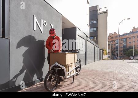 Junger Kurier mit roter Kleidung und Helm, der auf einem Lastenrad unterwegs ist, kommt am Versandort an, um ein Paket an eine Adresse in der Stadt zu liefern Stockfoto