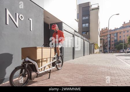 Junger Kurier mit roter Kleidung und Helm, der auf einem Lastenrad unterwegs ist, kommt am Versandort an, um ein Paket an eine Adresse in der Stadt zu liefern Stockfoto