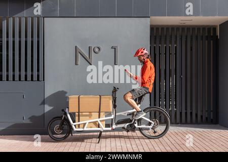 Junger Kurier mit roter Kleidung und Helm, der auf einem Lastenrad unterwegs ist, kommt am Versandort an, um ein Paket an eine Adresse in der Stadt zu liefern Stockfoto