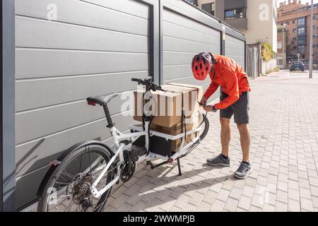 Junger Kurier mit roter Kleidung und Helm, der auf einem Lastenrad unterwegs ist, kommt am Versandort an, um ein Paket an eine Adresse in der Stadt zu liefern Stockfoto