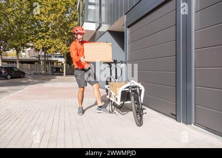 Junger Kurier mit roter Kleidung und Helm, der auf einem Lastenrad unterwegs ist, kommt am Versandort an, um ein Paket an eine Adresse in der Stadt zu liefern Stockfoto