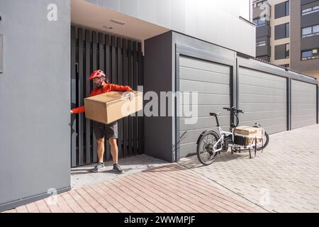 Junger Kurier mit roter Kleidung und Helm, der auf einem Lastenrad unterwegs ist, kommt am Versandort an, um ein Paket an eine Adresse in der Stadt zu liefern Stockfoto