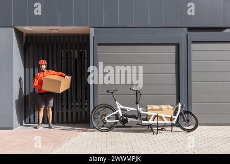 Junger Kurier mit roter Kleidung und Helm, der auf einem Lastenrad unterwegs ist, kommt am Versandort an, um ein Paket an eine Adresse in der Stadt zu liefern Stockfoto