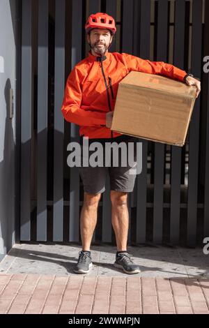 Junger Kurier mit roter Kleidung und Helm, der auf einem Lastenrad unterwegs ist, kommt am Versandort an, um ein Paket an eine Adresse in der Stadt zu liefern Stockfoto