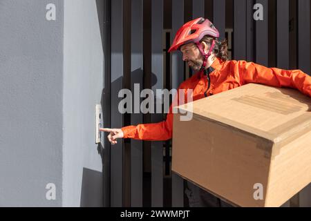 Junger Kurier mit roter Kleidung und Helm auf einem Lastenrad, der am Versandort ankommt, um ein Paket an eine Adresse in der Stadt zu liefern, Stockfoto