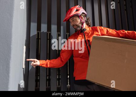 Junger Kurier mit roter Kleidung und Helm auf einem Lastenrad, der am Versandort ankommt, um ein Paket an eine Adresse in der Stadt zu liefern, Stockfoto