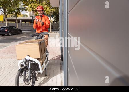 Junger Kurier mit roter Kleidung und Helm auf einem Lastenrad, der am Versandort ankommt, um ein Paket an eine Adresse in der Stadt zu liefern, Stockfoto