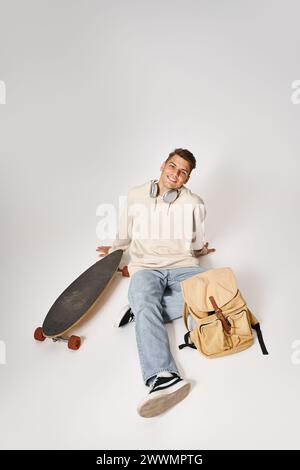 Attraktiver Schüler in Kopfhörern und lässigem Outfit, der mit Rucksack und Skateboard sitzt Stockfoto