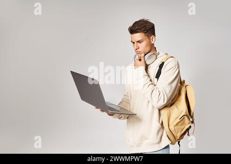 Durchdachter Schüler in Kopfhörern mit Rucksack-Netzwerkverbindung zum Laptop vor grauem Hintergrund Stockfoto