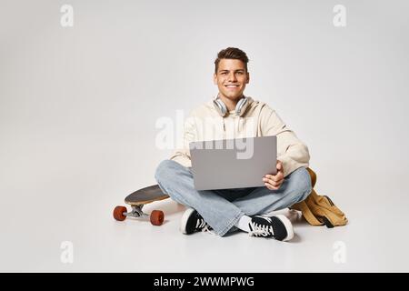 Lächelnder junger Schüler, der Kopfhörer mit Laptop verbindet und mit Rucksack und Skateboard sitzt Stockfoto