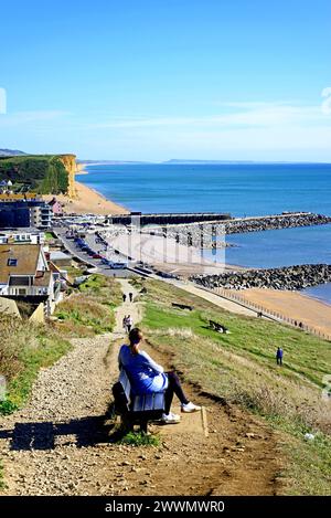 Erhöhter Blick über die Dächer der Stadt in Richtung Strand, Klippen und Landschaft mit einer Frau, die auf einer Bank im Vordergrund sitzt, West Bay, Großbritannien. Stockfoto