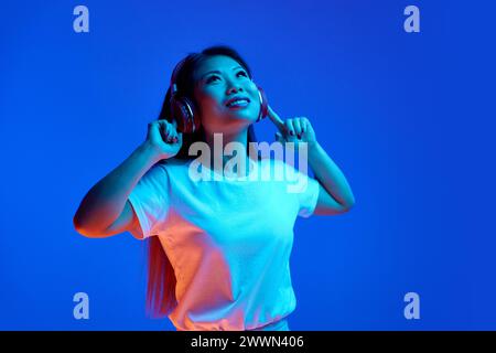 Porträt einer jungen attraktiven asiatischen Frau, die ihre Lieblingsmusik in Kopfhörern vor blauem Hintergrund im Neonlicht hört. Stockfoto